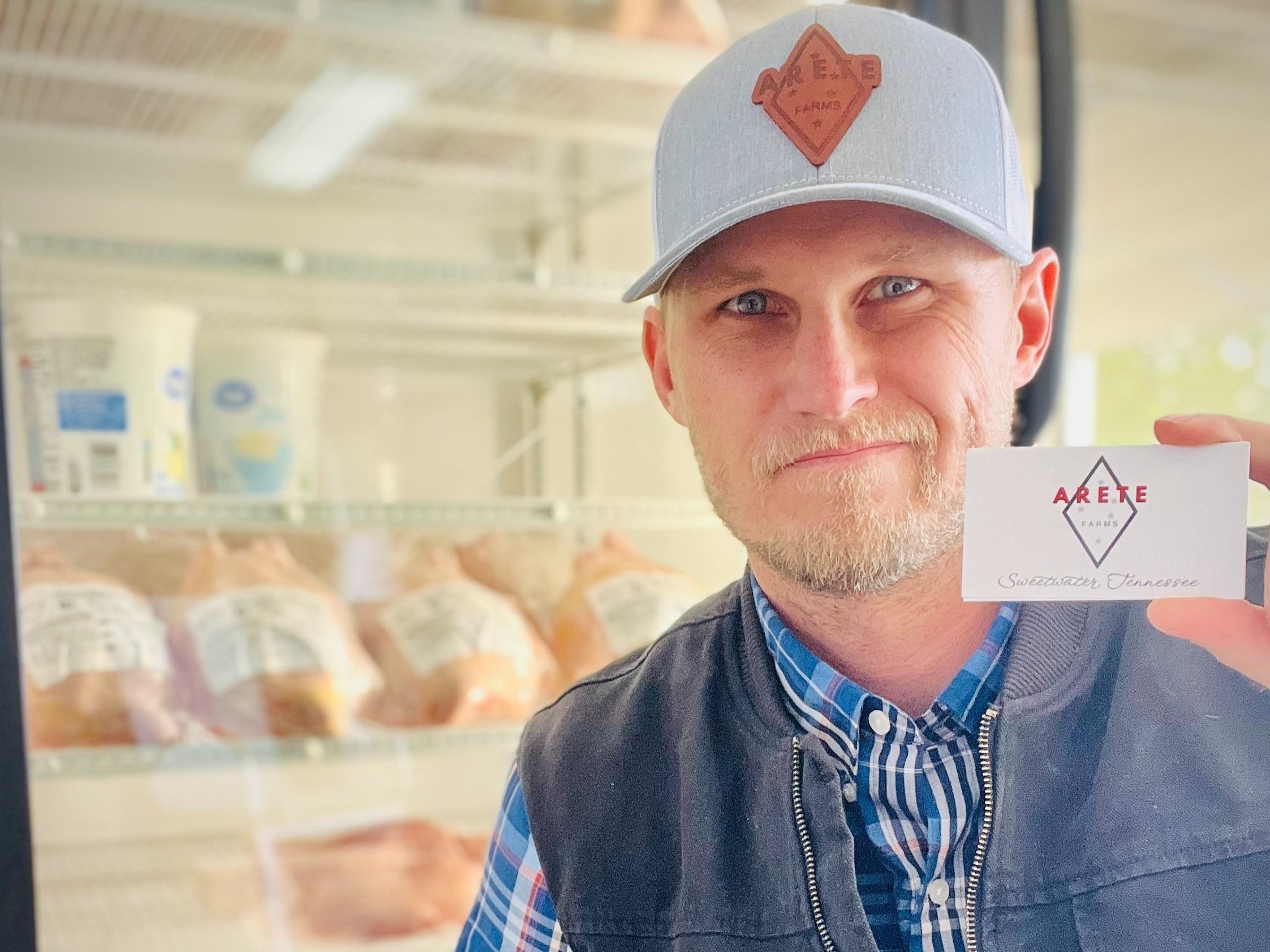 Man holding a business card for Arete Farms in front of a display case with packaged meats, showcasing local farm products in Sweetwater, Tennessee.